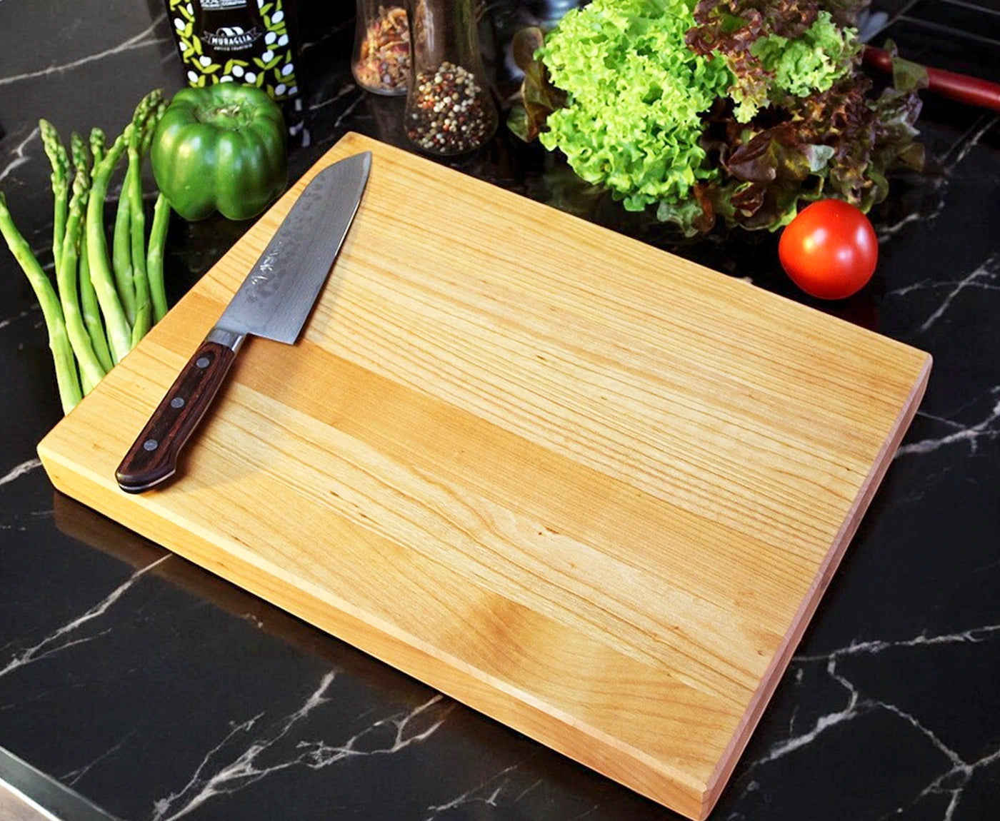 Küchenbrett Holz aus Kirschbaum mit Messer und Gemüse auf schwarzer Küchenarbeitsplatte.